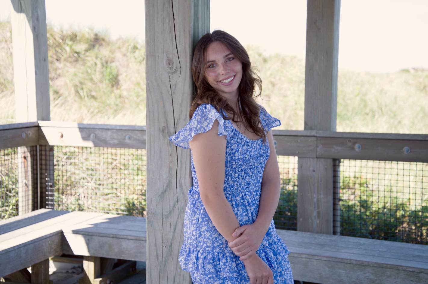 Senior portrait of young woman in blue floral dress leaning on beach boardwalk post