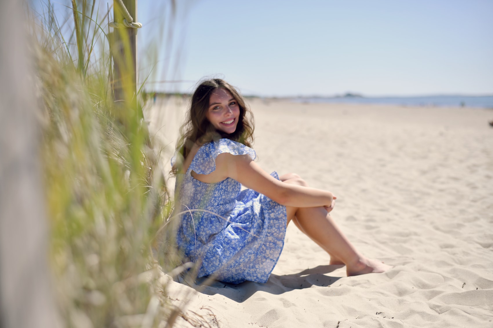 Senior portrait of young woman in blue floral dress sitting on beach smiling