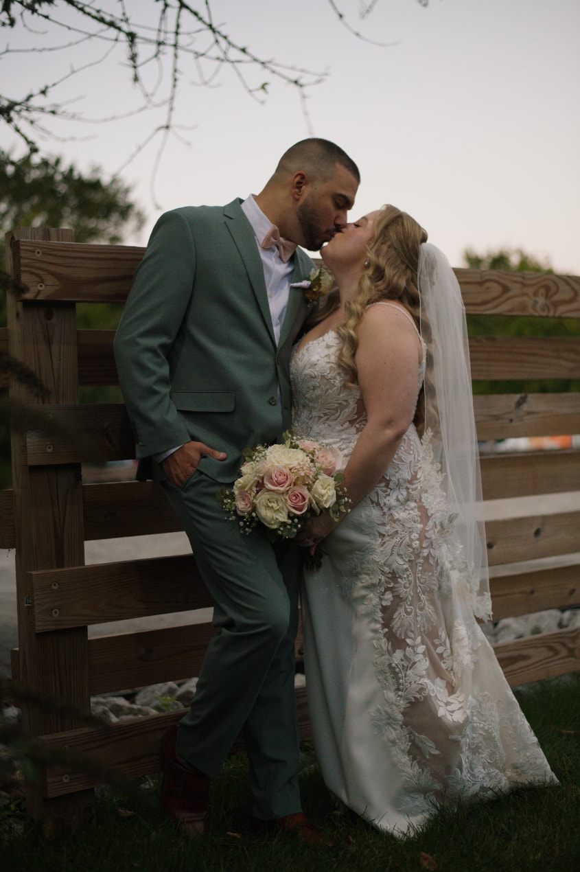 Bride and groom kissing against rustic wooden fence at dusk