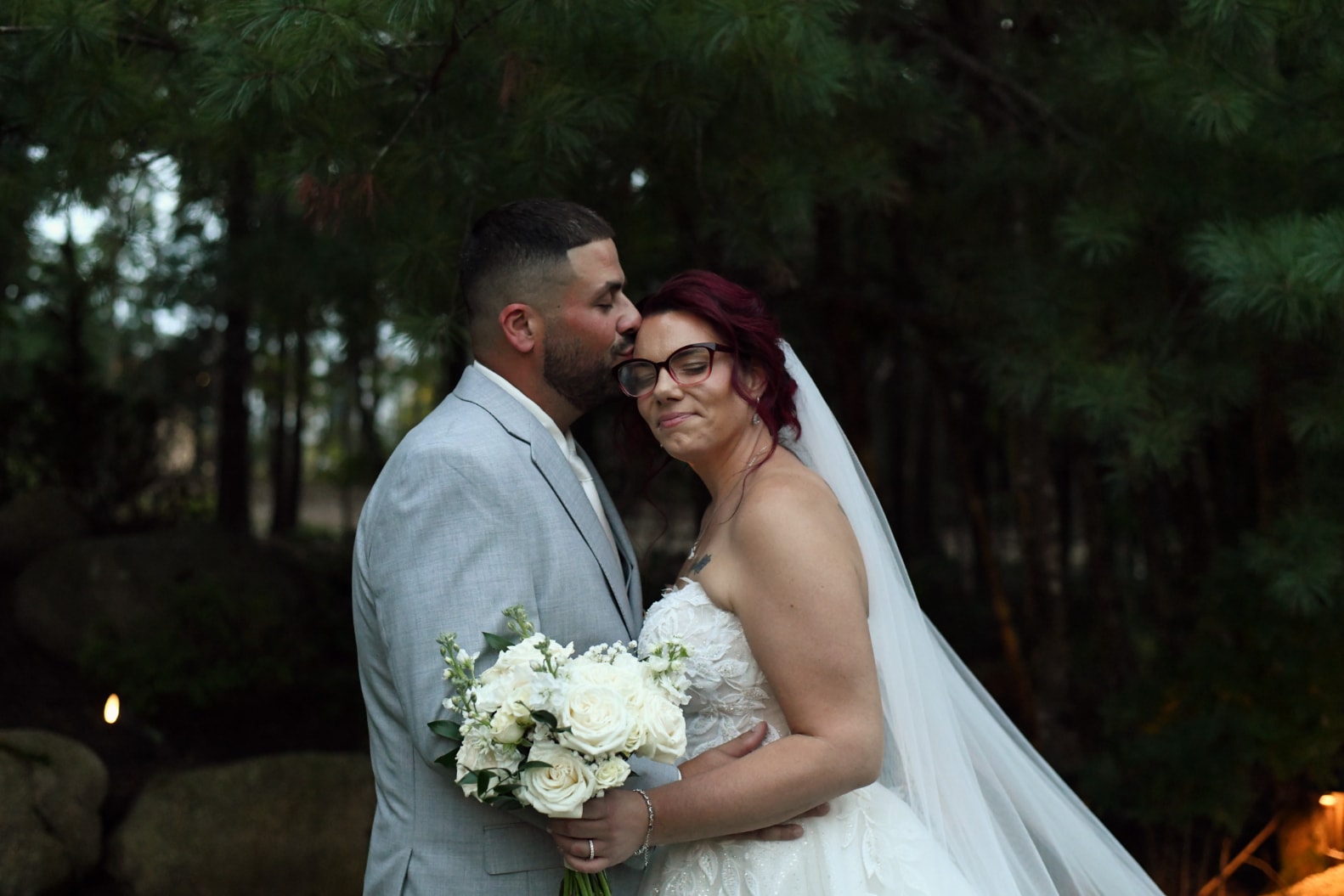 Groom kissing bride on cheek outdoors at dusk with white bouquet